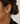 A close-up of a model's ear wearing a polished gold huggie hoop earring, with her hair in a bun and a black top visible against a light background.
