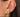 A close-up of a person's ear showcasing a curated stack of gold jewelry: a gold ear cuff with small clear stones, a gold huggie hoop with a green rectangular gemstone, a gold stud with a green heart-shaped gemstone, and a gold drop earring with a green oval gemstone.