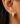 A close-up of a person's ear wearing a small gold huggie hoop earring adorned with pavé-set clear stones.