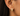 A close-up of a person's ear wearing a small gold huggie hoop earring adorned with pavé-set clear stones.