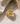 A gold ring with a double-loop knot design, featuring pavé-set clear stones on the top curve, displayed on a white marble surface with draped satin fabric.