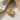 A gold ring with a double-loop knot design, featuring pavé-set clear stones on the top curve, displayed on a white marble surface with draped satin fabric.