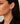 A close-up side profile of a woman wearing a small gold huggie hoop earring, with her dark hair and black blazer visible against a soft neutral background.