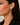 A close-up side profile of a woman wearing a small gold huggie hoop earring, with her dark hair and black blazer visible against a soft neutral background.