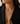 A close-up of a woman wearing a delicate gold necklace with three small letter charms spelling out 'MAYA' across the chest, styled with a black blazer.