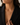 A close-up of a woman wearing a delicate gold necklace with three small letter charms spelling out 'MAYA' across the chest, styled with a black blazer.