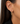 A close-up of a model's ear wearing a pair of delicate gold hoop earrings adorned with small clear stones, set against dark brown hair and a black blazer.