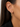 A close-up of a model's ear wearing a pair of delicate gold hoop earrings adorned with small clear stones, set against dark brown hair and a black blazer.