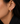 A close-up of a model's ear wearing a small gold huggie hoop earring with a clear marquise-cut center stone and a small peach accent.