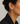 A close-up of a model's ear wearing a delicate gold T-shaped ear cuff adorned with small clear stones and a dangling bead drop, paired with a black blazer against a light gray background.