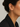 A close-up of a model's ear wearing a delicate gold T-shaped ear cuff adorned with small clear stones and a dangling bead drop, paired with a black blazer against a light gray background.