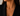A close-up of a model wearing a delicate gold necklace with the word 'MAMA' spelled out in small letter pendants, styled with a black blazer.
