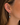 A close-up of a model's ear wearing a gold hoop earring in the cartilage and a matching drop earring with a row of clear stones and a dark gray oval pendant.