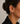 A close-up side profile of a woman wearing a delicate gold ear cuff adorned with small clear stones and a dangling teardrop charm, paired with a black collared shirt against a neutral gray background.