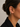 A close-up side profile of a woman wearing a delicate gold ear cuff adorned with small clear stones and a dangling teardrop charm, paired with a black collared shirt against a neutral gray background.