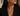 A close-up of a woman wearing a gold chain necklace with a spherical pendant, one side of which is encrusted with small clear stones. She is wearing a black blazer and a matching gold and stone stud earring.