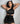 A woman posing in a black satin halter-neck mini dress with cutouts, set against a plain light gray studio background.