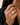 A close-up of a person's hand wearing a gold ring with a large, multi-faceted clear baguette-cut gemstone, set against a dark blazer and neutral background.